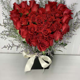 Heart-shaped arrangement of red roses in a black box with purple filler flowers and a white bow.