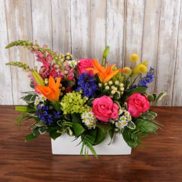 Low rectangular arrangement of bright pink roses, orange lilies, blue flowers, and mixed blooms in a white container