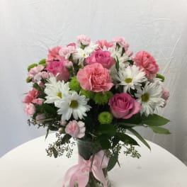 Pink roses, carnations, and white daisies in a glass vase with a pink ribbon.