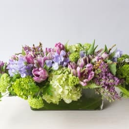 Low floral arrangement with purple, pink, white, and green blooms in a rectangular vase