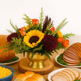 Low centerpiece with a yellow sunflower, orange roses, and burgundy mums in a gold vase on a dinner table.