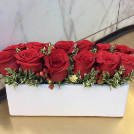 Red roses arranged in a white rectangular vase with small variegated greenery