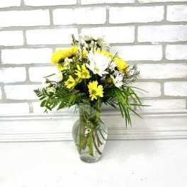 Small bouquet of yellow and white daisies arranged with greenery in a clear glass vase