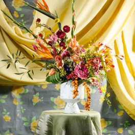 Tall modern arrangement of tropical and mixed flowers in a white pedestal vase on a draped table.