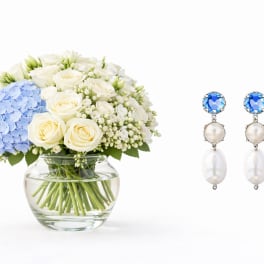 White roses and blue hydrangea in a glass vase beside blue-and-pearl earrings