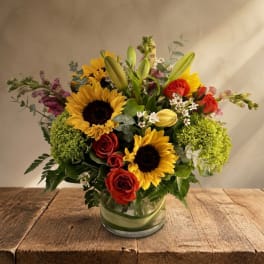 Mixed bouquet with sunflowers, red roses, lilies, and green blooms in a glass vase