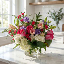 Mixed bouquet of pink roses, lilies, purple blooms, and white hydrangeas in a glass vase