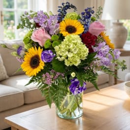 Mixed bouquet with sunflowers, roses, hydrangea, and purple blooms in a glass vase