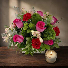 Pink roses and red carnations arranged in a low square vase with a candle nearby.