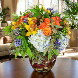 Mixed bouquet of orange roses, yellow orchids, and blue hydrangeas in a glass vase