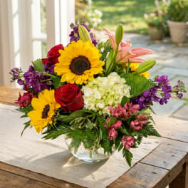 Mixed bouquet in a glass vase with sunflowers, roses, lilies, and purple blooms