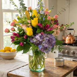 Mixed bouquet of yellow roses, purple hydrangea, and pink blooms in a glass vase