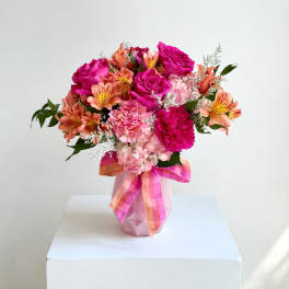 Pink and orange mixed flower bouquet in a pink vase with a ribbon
