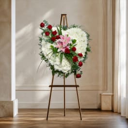 Heart-shaped floral wreath with white blooms, red roses, and pink lilies on an easel