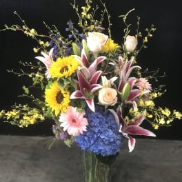 Tall mixed bouquet in a glass vase with sunflowers, lilies, roses, and blue hydrangea