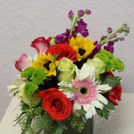 Mixed bouquet of red, yellow, white, and purple flowers in a square glass vase