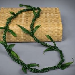 Braided green leaf lei draped over a tan woven mat against a gray background