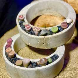 Stacked concrete ring planters filled with small multicolored succulent stones on black gravel.