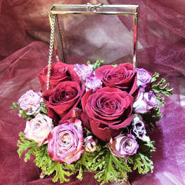 Compact arrangement of red and lavender roses in a gold metal and glass box on a burgundy fabric background