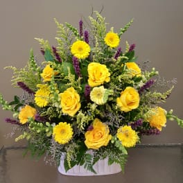 Large yellow rose and gerbera daisy arrangement with purple accents in a white basket.