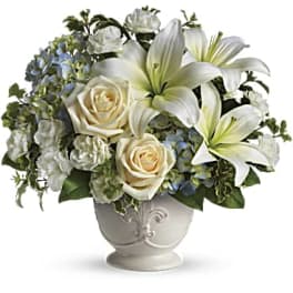 White lilies, roses, carnations, and blue hydrangeas arranged in a white ceramic pedestal vase