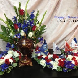 Floral funeral tribute with red, white, and blue flowers around an urn and small flags.