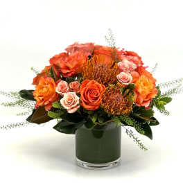 Low orange and peach rose arrangement with pincushion protea in a glass cylinder vase.