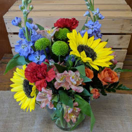 Mixed bouquet with sunflowers, roses, carnations, and blue flowers in a glass vase