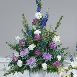 Tall fan-shaped arrangement of purple roses, lavender mums, and white blooms in a white urn vase