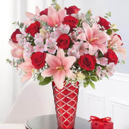 Pink lilies and red roses in a red vase with a small gift box nearby