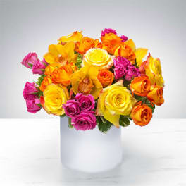 Bright arrangement of yellow, orange, and hot pink roses with orchids in a white cylinder vase