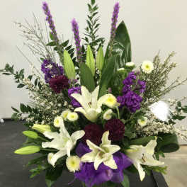 Tall floral arrangement with white lilies, purple blooms, and a purple ribbon in a vase