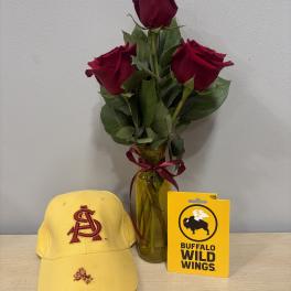Three red roses in a glass vase with a ribbon, beside a yellow cap and gift card.
