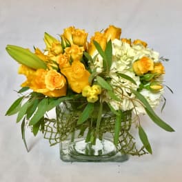 Low arrangement of yellow roses, lily buds, and white hydrangea in a clear glass cube vase