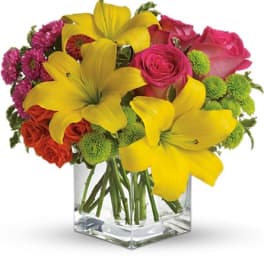 Bouquet of yellow lilies, pink roses, and green button mums in a clear square vase