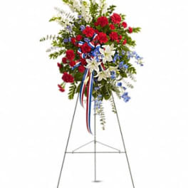 Patriotic standing spray of red carnations, white lilies, and blue flowers with ribbon on a metal easel