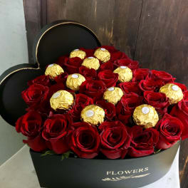 Red roses arranged with gold-wrapped chocolates in a black heart-shaped box