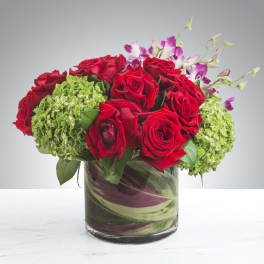 Low arrangement of red roses, green hydrangeas, and purple orchids in a clear glass cylinder vase