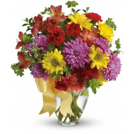 Bright mixed bouquet of yellow daisies, purple mums, and red carnations in a clear vase with yellow ribbon
