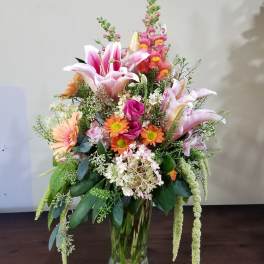 Tall mixed arrangement of pink lilies, orange daisies, and assorted flowers in a clear glass vase