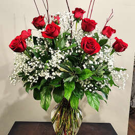 Tall arrangement of red roses and white filler flowers in a clear glass vase on a dark table