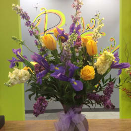 Tall bouquet of yellow tulips, purple irises, and lilac in a vase with a purple bow