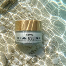 A cream jar with a wood lid sits on pebbles under rippling water.