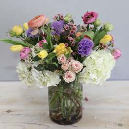 Mixed arrangement with white hydrangeas, pink spray roses, yellow tulips, and purple blooms in a clear glass vase