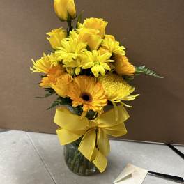 Yellow roses and gerbera daisies in a glass vase with a yellow ribbon