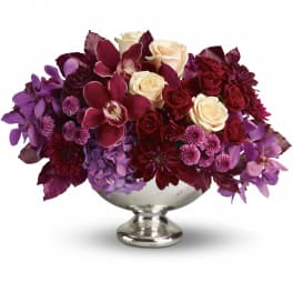Low centerpiece of ivory roses, burgundy blooms, and purple orchids in a silver pedestal bowl