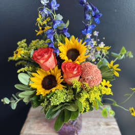 Bouquet with sunflowers, coral roses, and blue delphinium in a glass vase