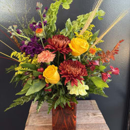 Mixed bouquet with yellow roses, burgundy mums, and purple flowers in an amber vase
