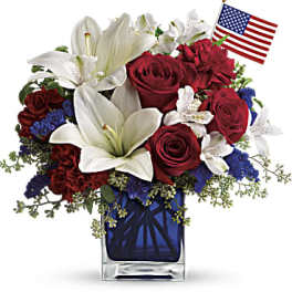 Patriotic arrangement with white lilies, red roses, and blue accents in a blue glass vase with a small US flag