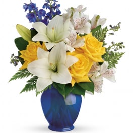Yellow roses and white lilies in a blue vase with blue delphinium